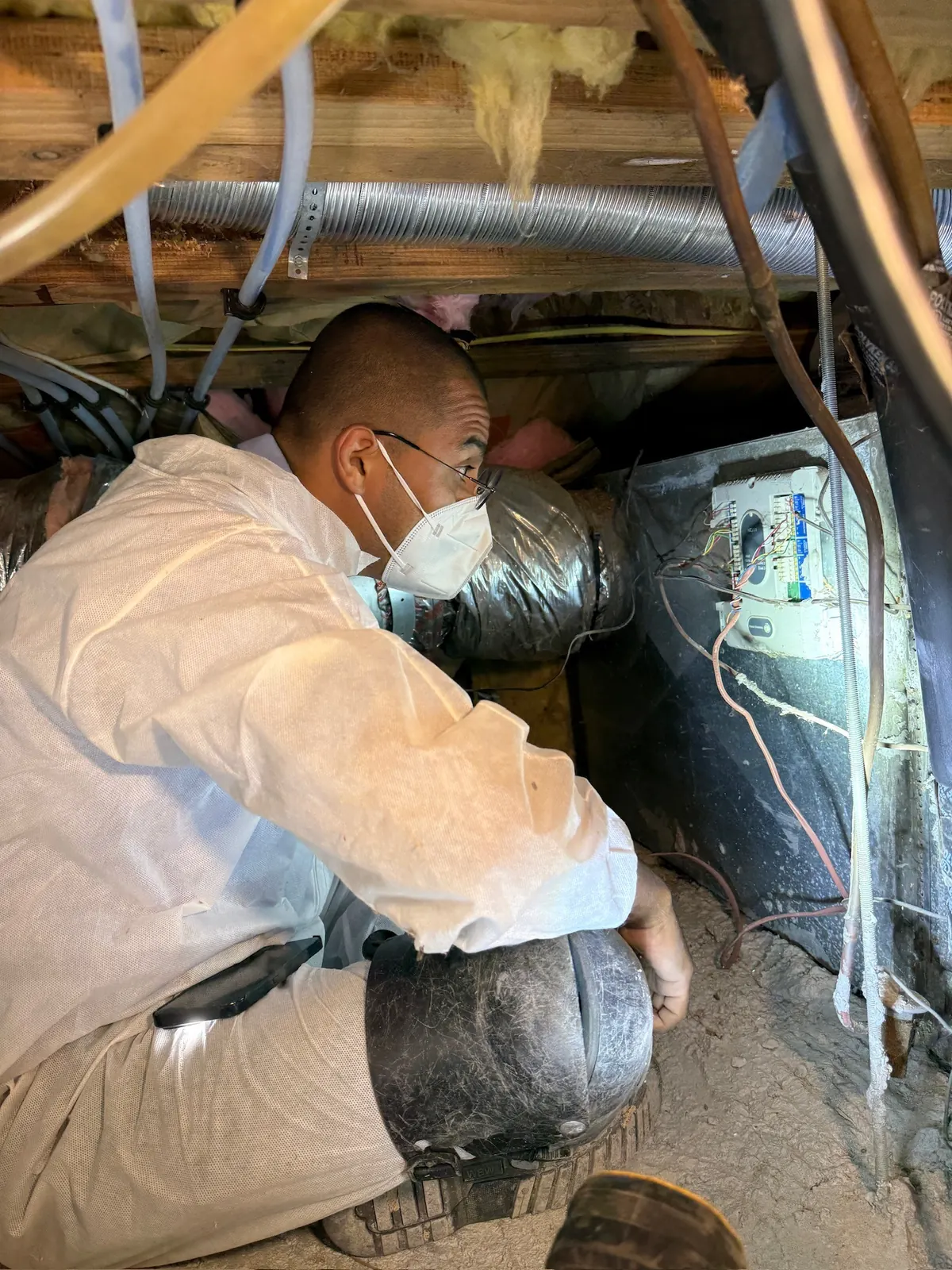 AirtekPros technician in coveralls and a respirator sealing flexible duct connections in a crawlspace