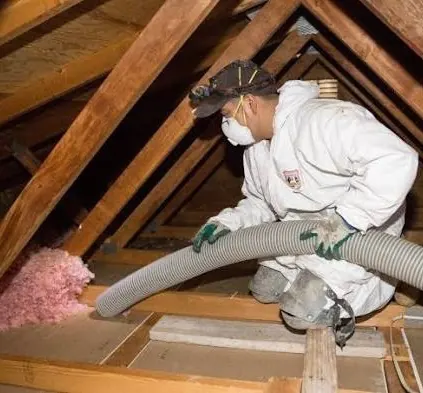 AirtekPros technician in protective coveralls and respirator removing attic insulation with a vacuum hose